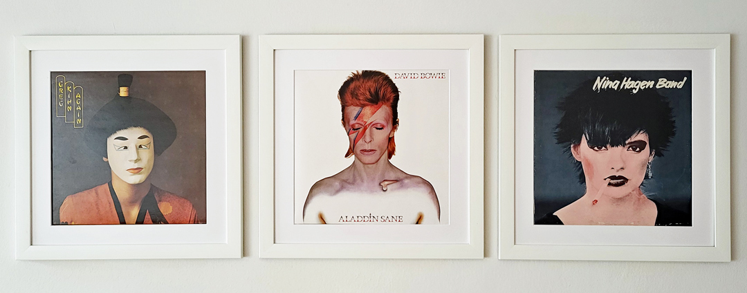 Drei Plattencover mit geschminkten Personen – darunter David Bowie und Nina Hagen – in weißen Holzrahmen präsentiert.