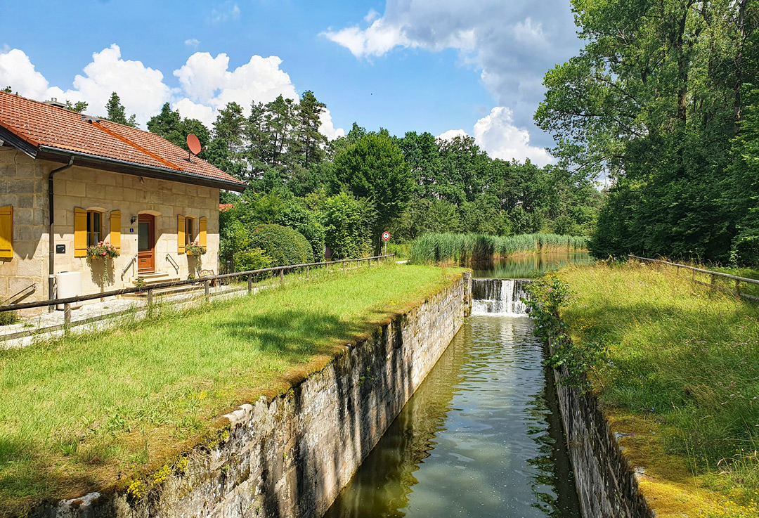 Schleuse am Ludwig-Donau-Main-Kanal, dem historischen Vorläufer des heutigen Main-Donau-Kanals. Zu sehen sind die alte Schleusenanlage mit begrünten Uferwänden, das ehemalige Schleusenwärterhäuschen aus Sandstein und Bäume unter einem blauen Himmel.