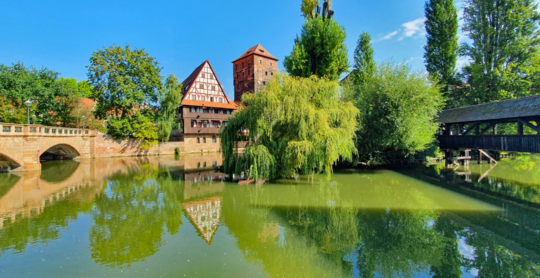 Blick auf die Pegnitz in Nürnberg mit der Maxbrücke links, dem Fachwerkbau des Weinstadels und dem Wasserturm in der Mitte, sowie dem Henkersteg als überdachte Holzbrücke rechts, umgeben von Bäumen und spiegelndem Wasser.