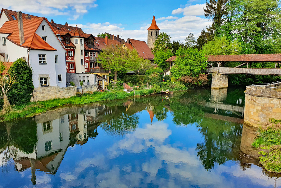 Blick auf die Altstadt von Lauf an der Pegnitz mit Fachwerkhäusern, überdachter Holzbrücke und dem Fluss, der die Gebäude malerisch spiegelt.