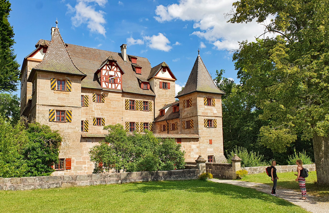 Das Petz'sche Schloss in Schwarzenbruck, ein historischer Sandsteinquaderbau aus dem 16. Jahrhundert, umgeben von Bäumen und einer Steinmauer, bei Sonnenschein und blauem Himmel.