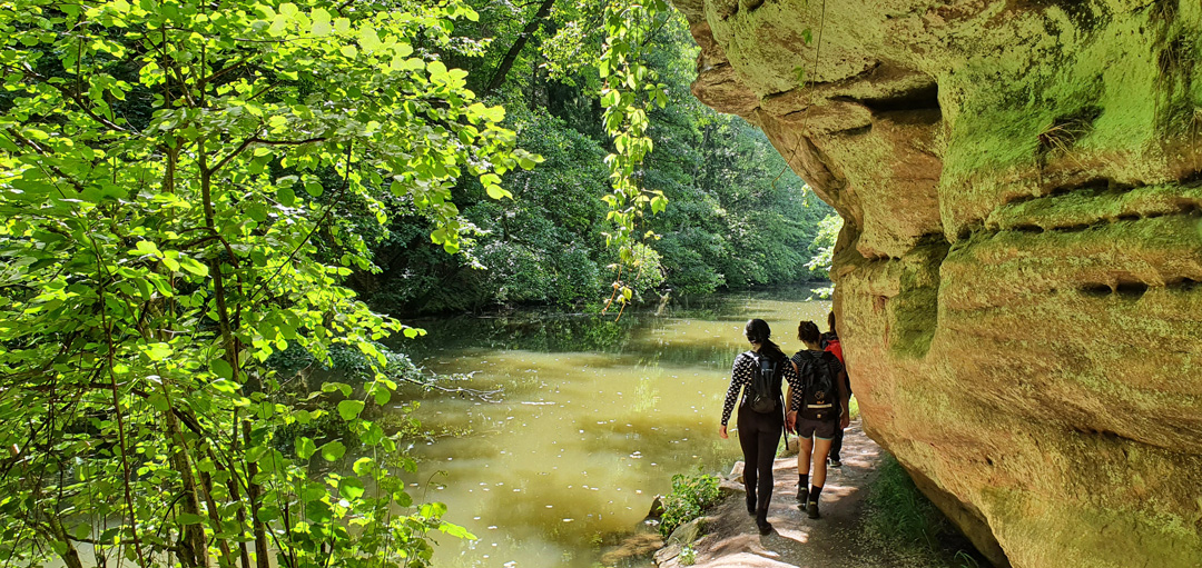 Drei Frauen auf einem Wanderpfad zwischen dem Flüsschen Schwarzach und einem imposanten, überhängenden Sandsteinfelsen.