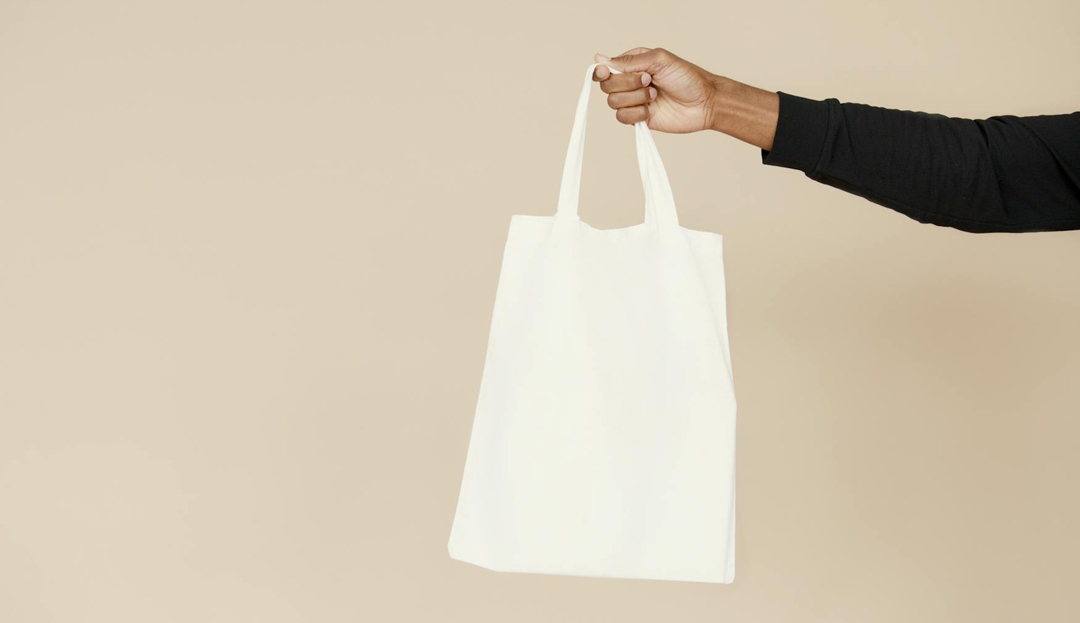 Hand hält einen weißen Jutebeutel vor beigem Hintergrund – Symbol für Less Waste, Wiederverwendung und abfallbewusstes Einkaufen.