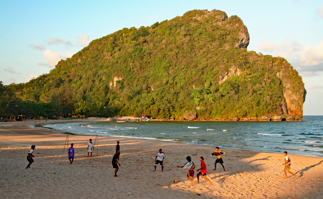 Junge Männer spielen am Strand in Thailand Fußball, während die Sonne Sand, Meer und Felsen in warmes Licht taucht.