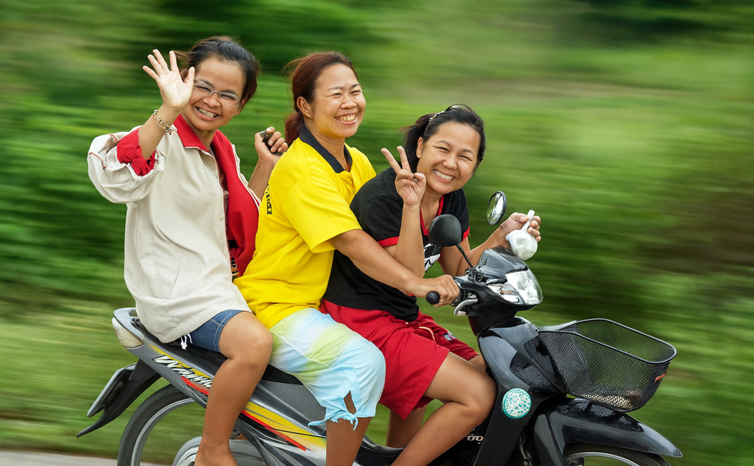 Drei junge Frauen fahren auf einem Motorrad in Thailand, lachen und winken in die Kamera – ein Moment voller Lebensfreude und Spontaneität.