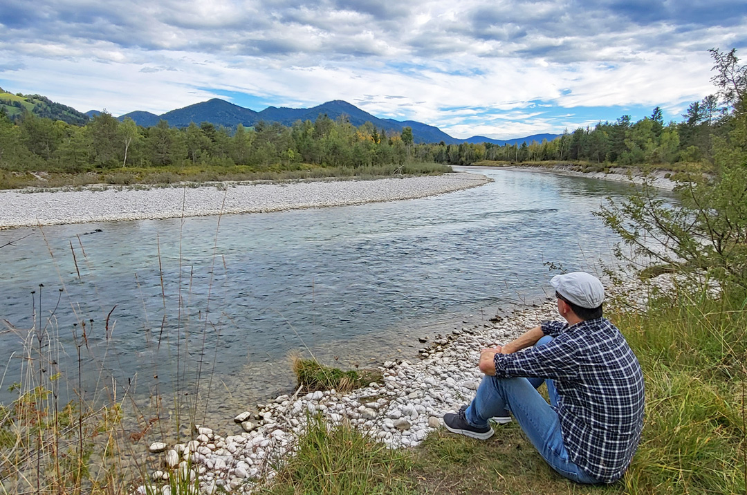 Autor Christof Herrmann sitzt am Ufer der Isar und blickt über den Fluss zu den Bergen.