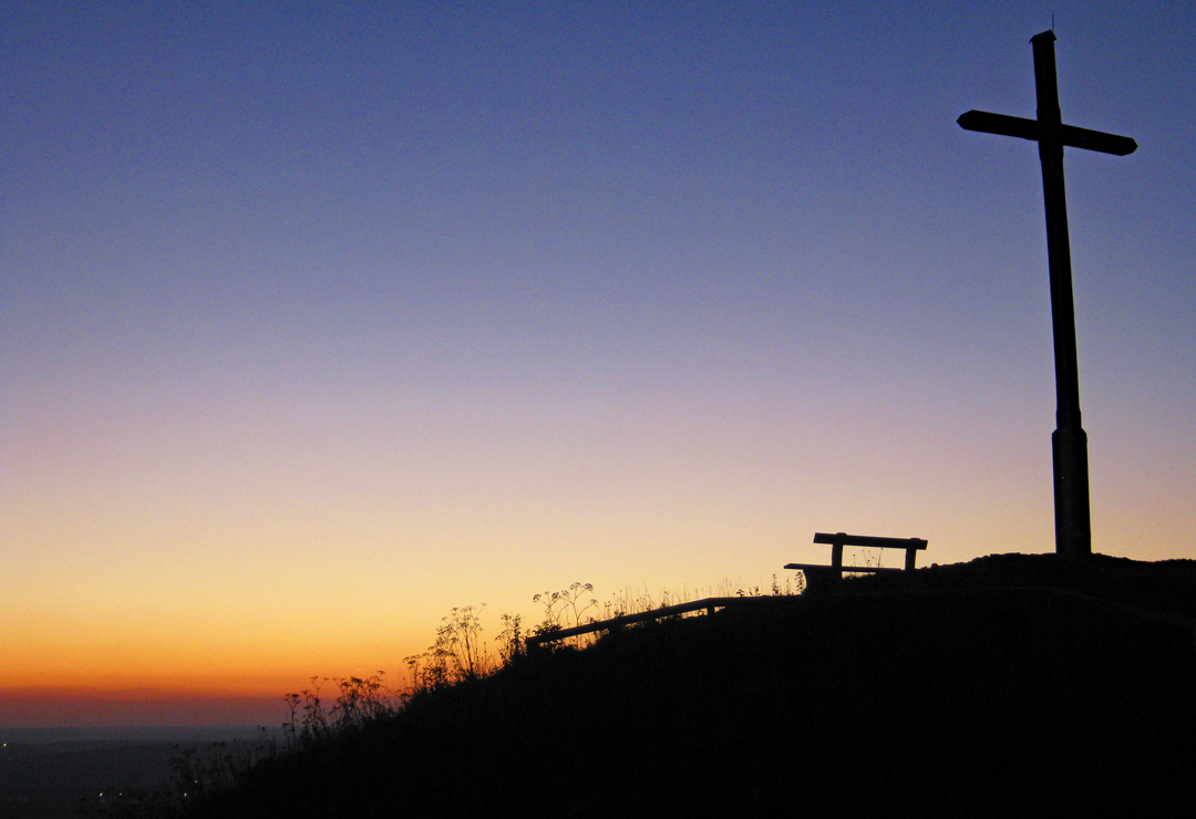 Grasiger Hang bei Sonnenuntergang mit Bank und Gipfelkreuz als Silhouetten.